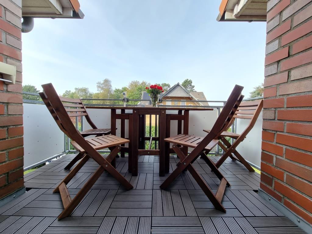 Terrasse mit Holztisch, vier Stühlen und Blumenarrangement. Blick auf grüne Umgebung.