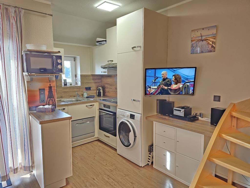 Küche mit Spüle, Herd und Waschmaschine. Fernseher an der Wand. Holztreppe rechts.