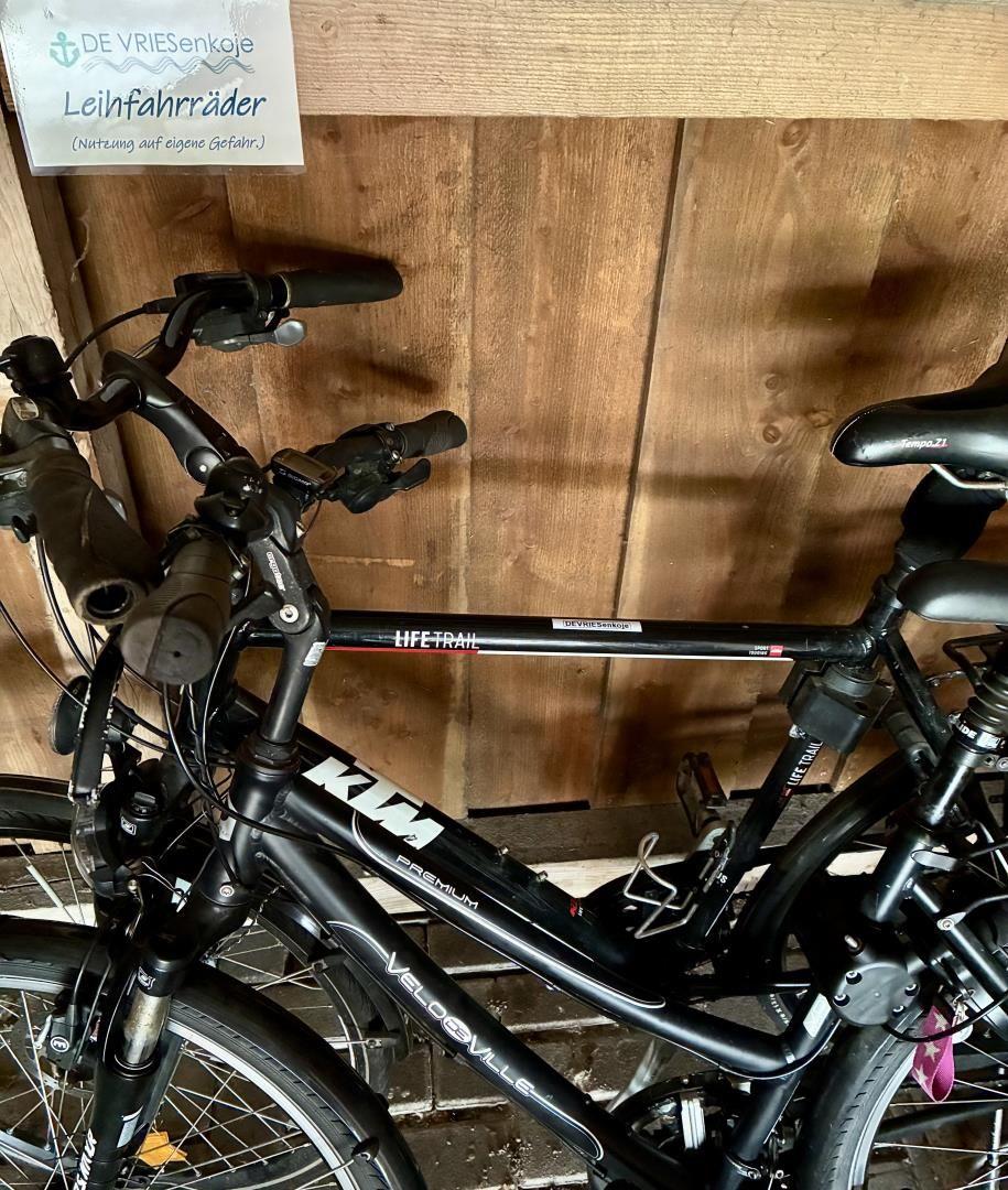 Zwei schwarze Fahrräder stehen in einer Holzwandgarage. Ein Schild zeigt Leihfahrräder an.