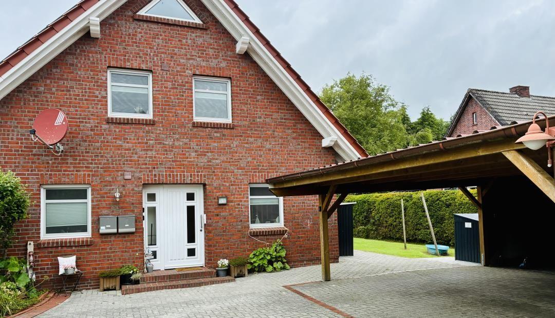 Rotes Backsteingebäude mit Eingangstür und Carport. Grünflächen und Satellitenschüssel sichtbar.