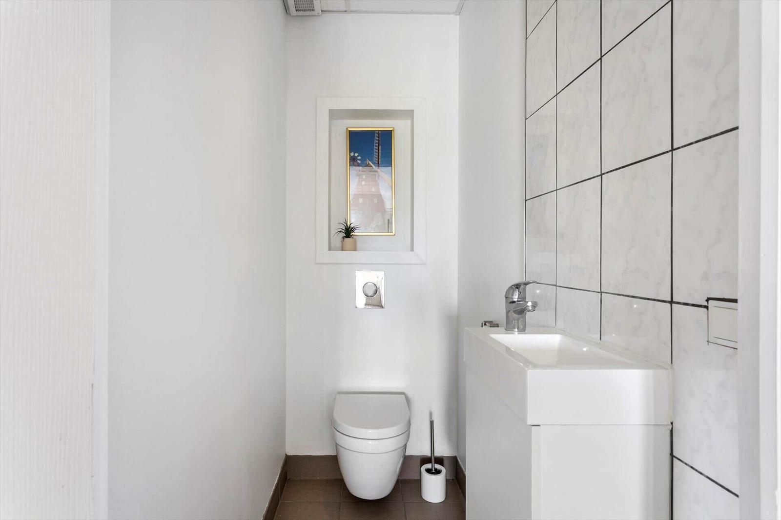 Bathroom with toilet, sink, and tiled wall. A picture and plant on the shelf.