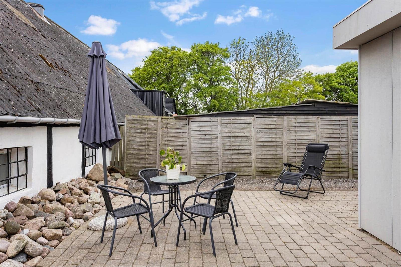 Terrace with table, chairs, and lounge chair. Backyard with wooden fence and trees.
