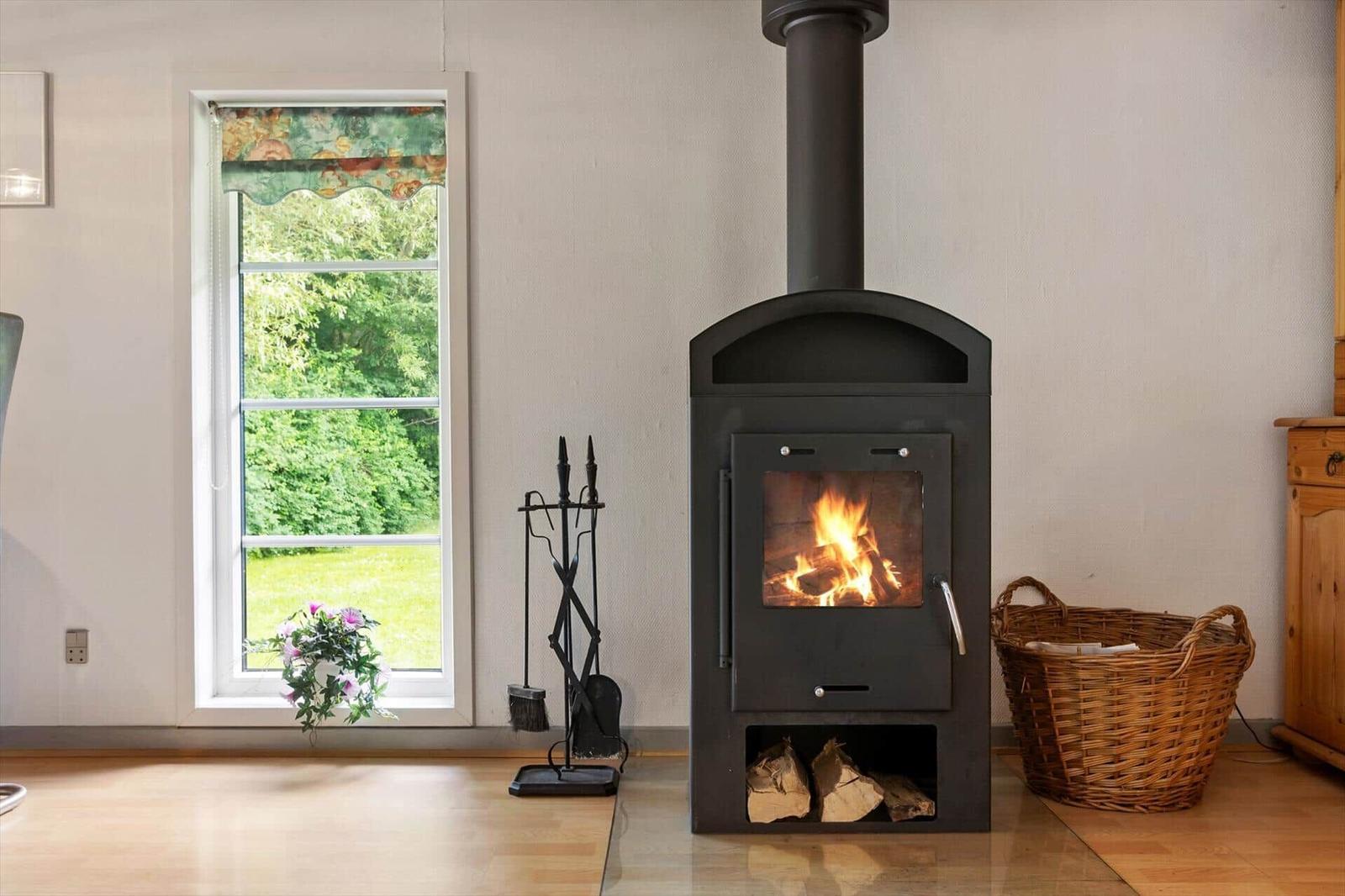 Wood-burning stove, window with view of greenery, basket, and fireplace tools.