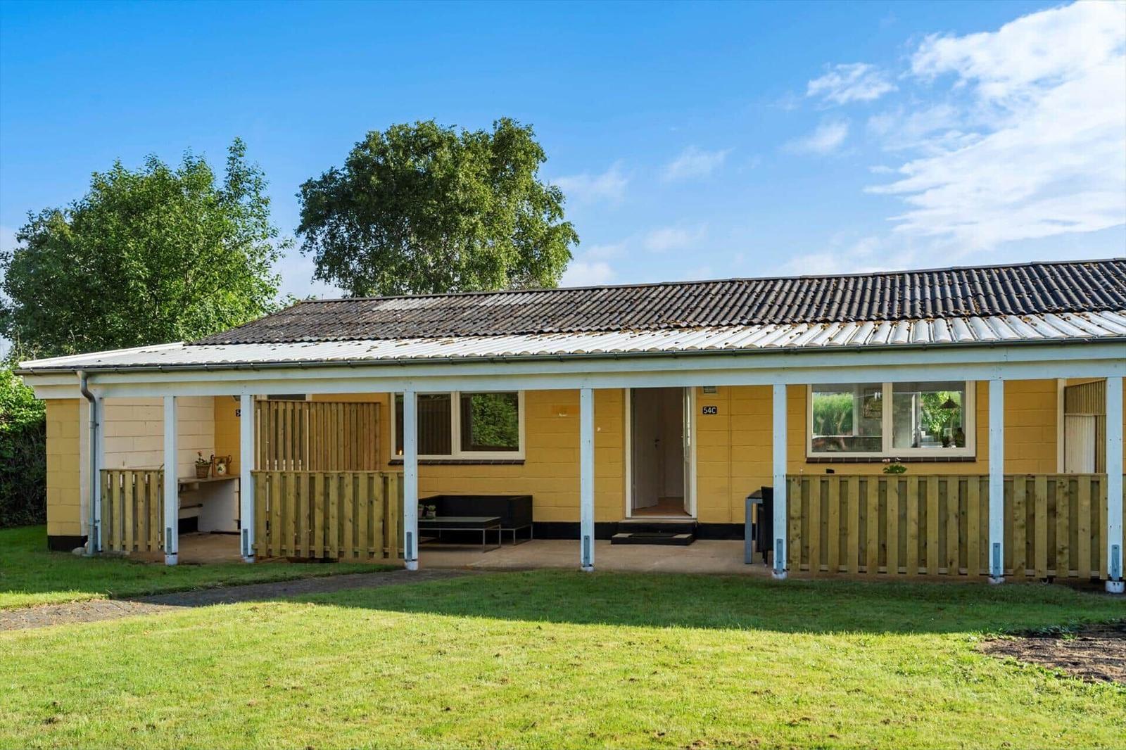 A yellow holiday home with a covered terrace and wooden railing. Background: trees and blue sky.