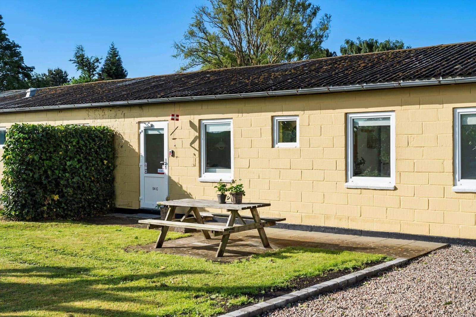 Yellow holiday house with wooden table, lawn, and windows. Backyard with hedge and door.