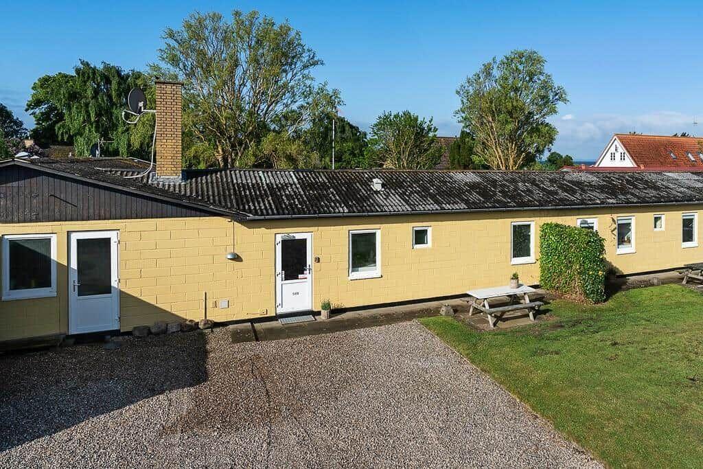Gelbes Ferienhaus mit mehreren Fenstern, Grillplatz und Rasenfläche. Hintergrund mit Bäumen und klarer blauer Himmel.