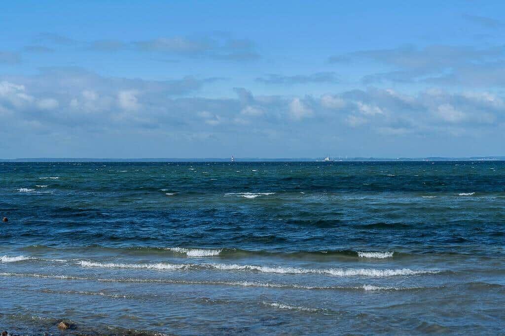 Meeresblick mit Wellen und Segelbooten im Hintergrund unter blauem Himmel.