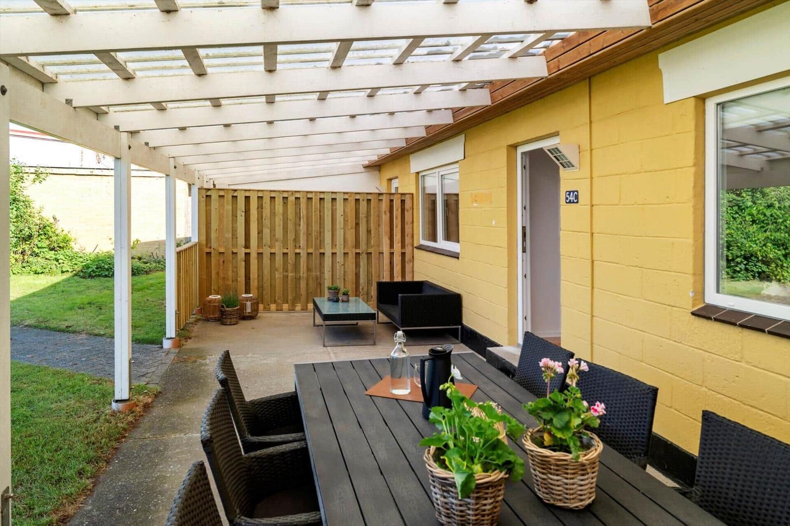 Terrasse mit Tisch, Stühlen und Pergola neben gelbem Haus