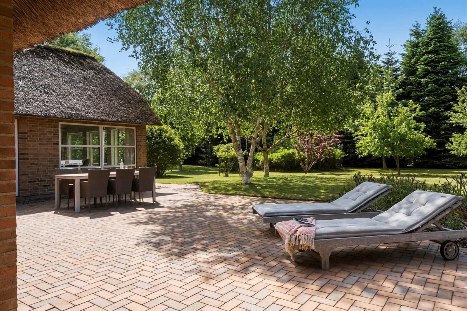 Terrasse mit Liegestühlen, Tisch und Stühlen. Hintergrund: Gras, Bäume und Gebäude mit Strohdach.