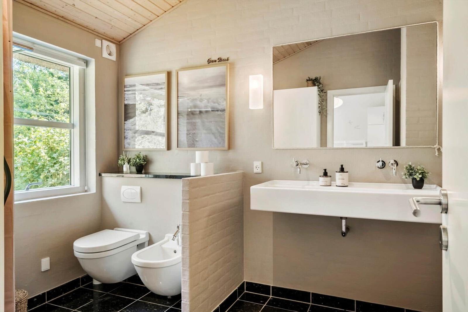Badezimmer mit Toilette, Bidet, Waschbecken und Spiegel. Fenster mit Blick auf Grün.