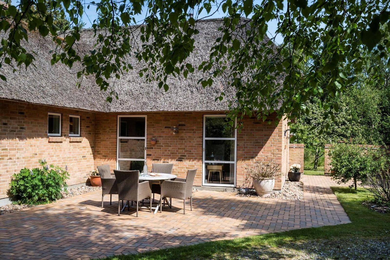 Haus mit Strohdach und Terrasse mit Tisch und Stühlen. Umgeben von Grün und Bäumen.