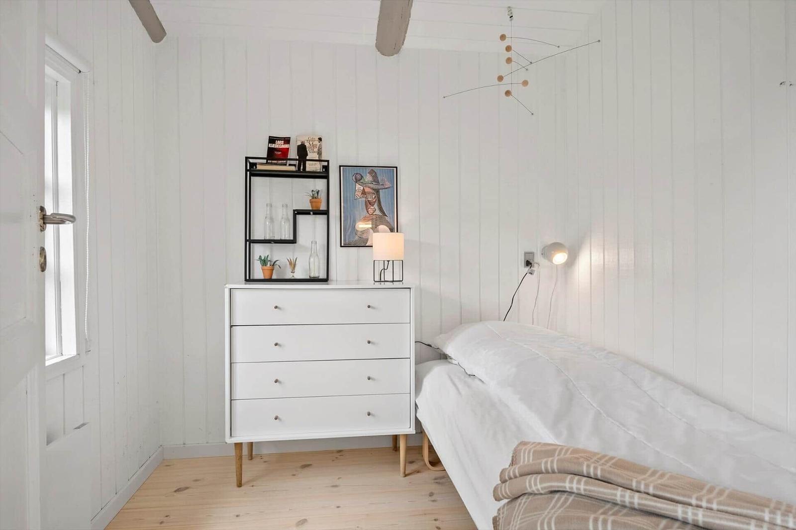 Bedroom with white dresser, bed, and wall shelf. Wooden floor and white walls.