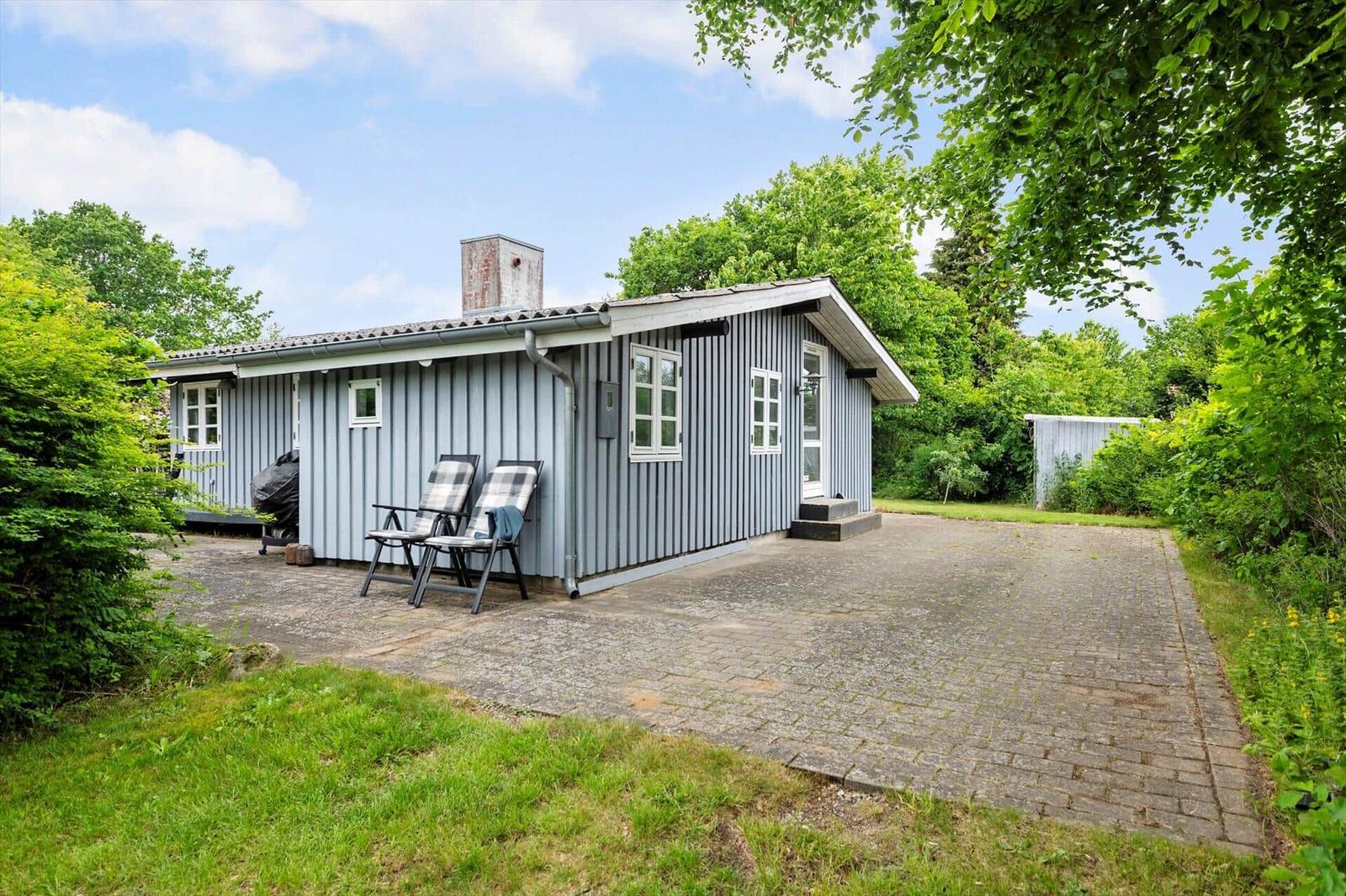 A small house with wooden siding, two chairs, and a grill on a paved patio.