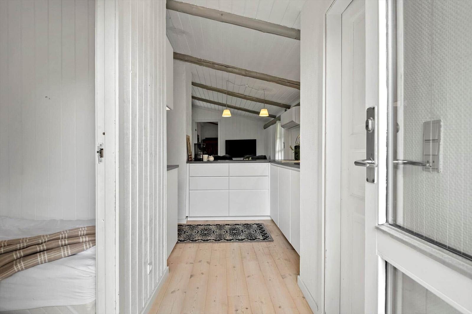 Bedroom with bed and door. Corridor to kitchen with white built-in cabinetry and wooden floor.