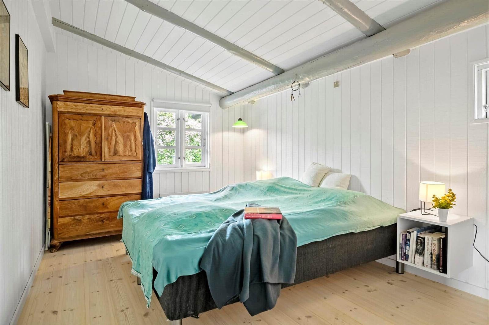 Bedroom with bed, wooden dresser, and nightstand. White walls and wooden floor.