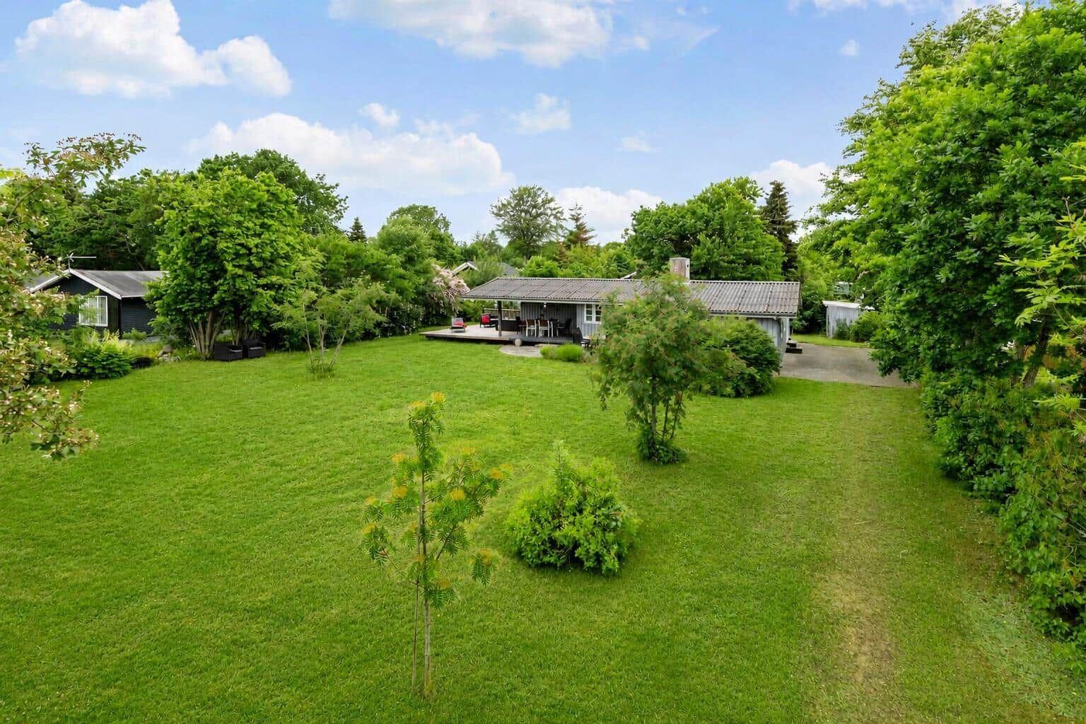 House with large garden, terrace, and grill area. Surrounded by trees under blue sky.