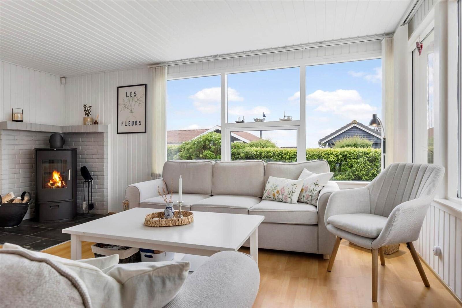 Wohnzimmer mit Sofa, Sessel und Kamin. Große Fenster bieten Blick auf Garten.