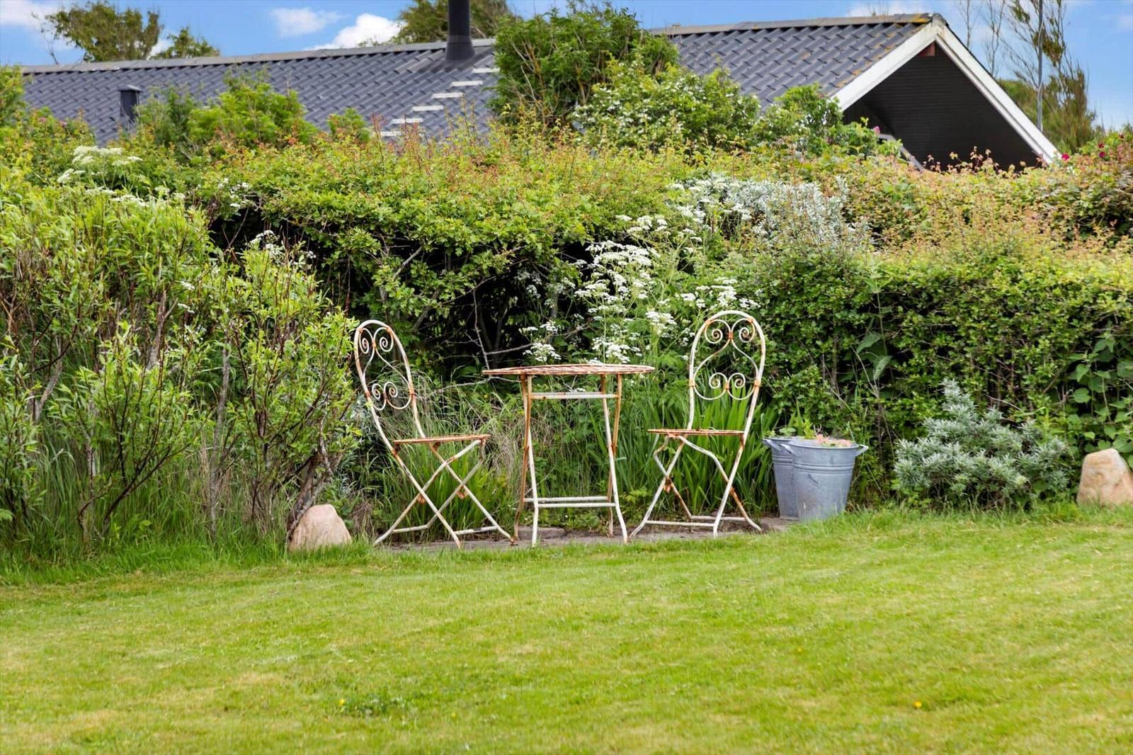 Garten mit Tisch und zwei Stühlen vor grüner Hecke und Haus.