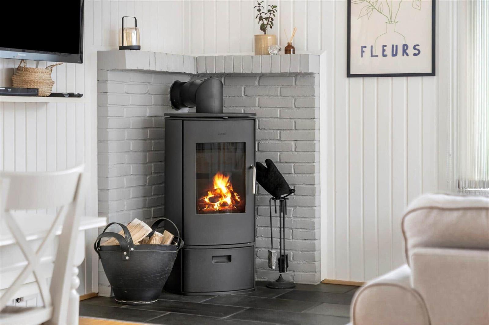 Ein Kaminofen mit brennendem Feuer steht in einem Raum mit weißer Wandverkleidung und einem Holzkorb mit Brennholz.