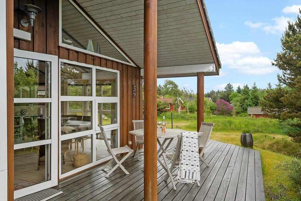 Deck with table and chairs, wooden wall, glass doors, view of meadow and trees.