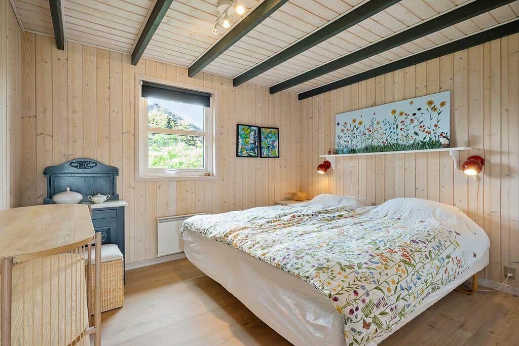 Bedroom with wooden walls, bed with floral cover, window and wall art.