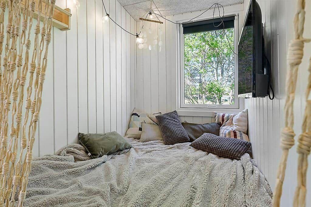 A room with mattress, pillows, and blanket against the wall. Window, TV, and hammock.
