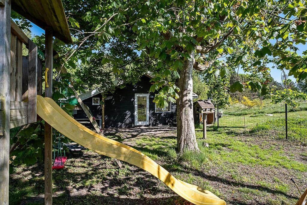 Black wooden house with playground and yellow slide under trees.