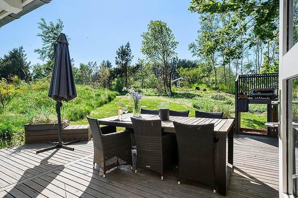 Deck with table, chairs, and umbrella. Green areas and trees in the background.