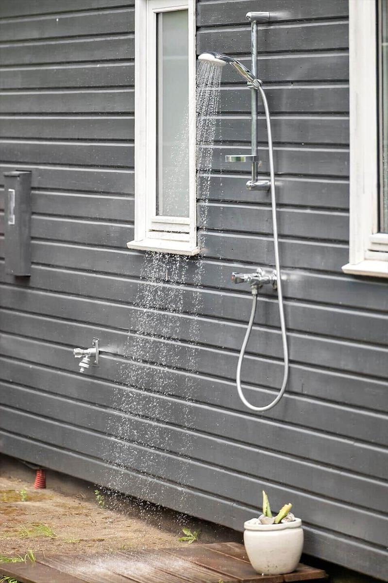 Outdoor shower on gray wooden house wall with water stream and plant in foreground.