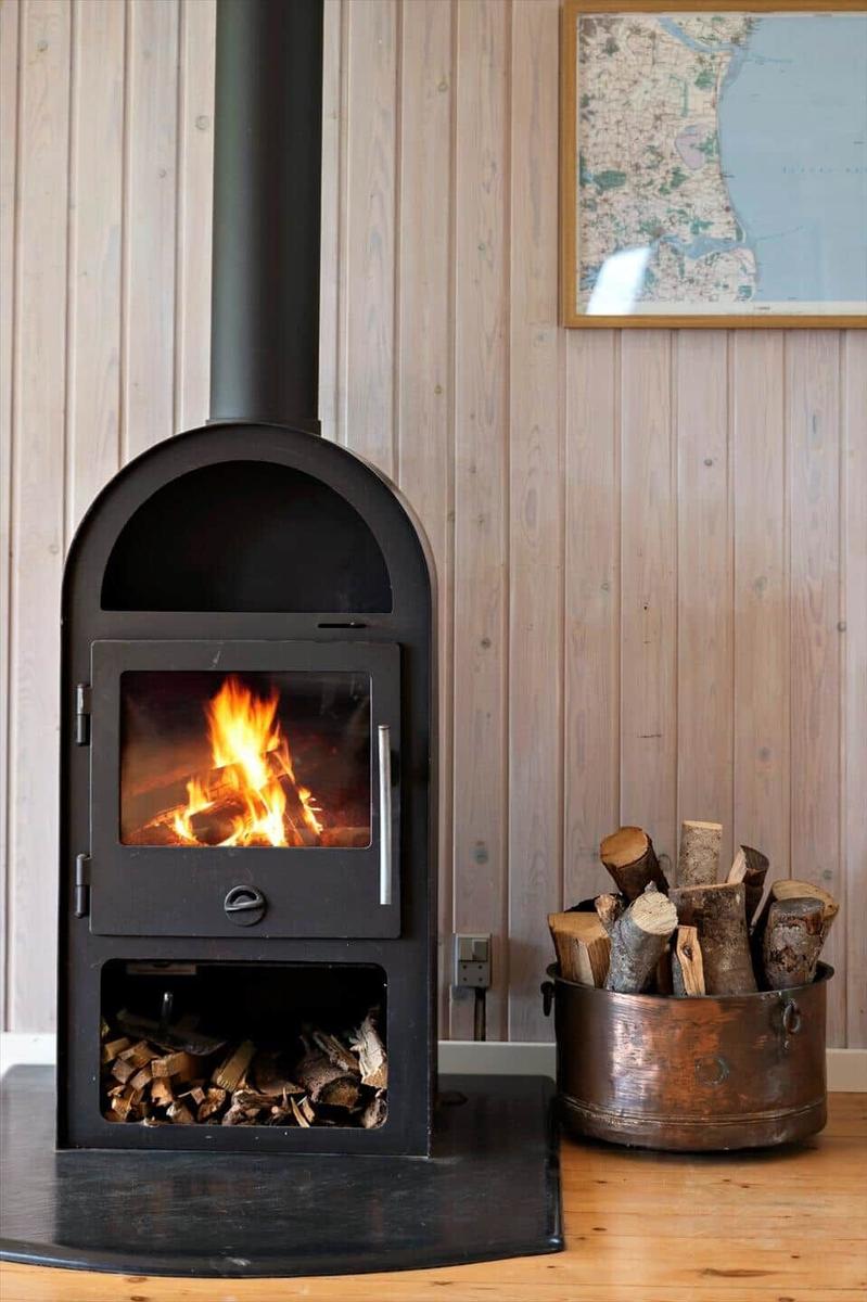 Wood stove with burning logs, beside bucket with firewood against wood wall.