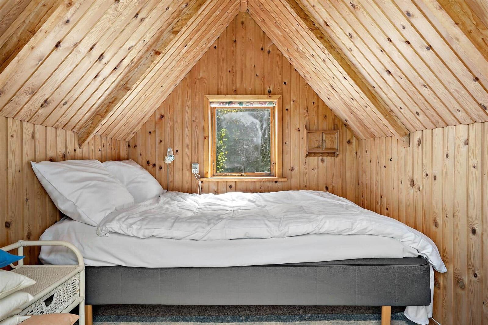 Attic room with wooden walls and ceiling, bed with white bedding and small window.