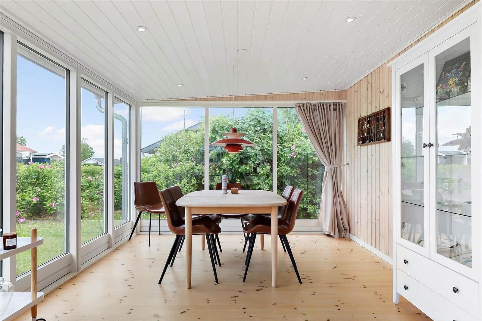 Dining room with wooden floor, glass walls, and brown chairs. Hanging lamp and glass cabinet.