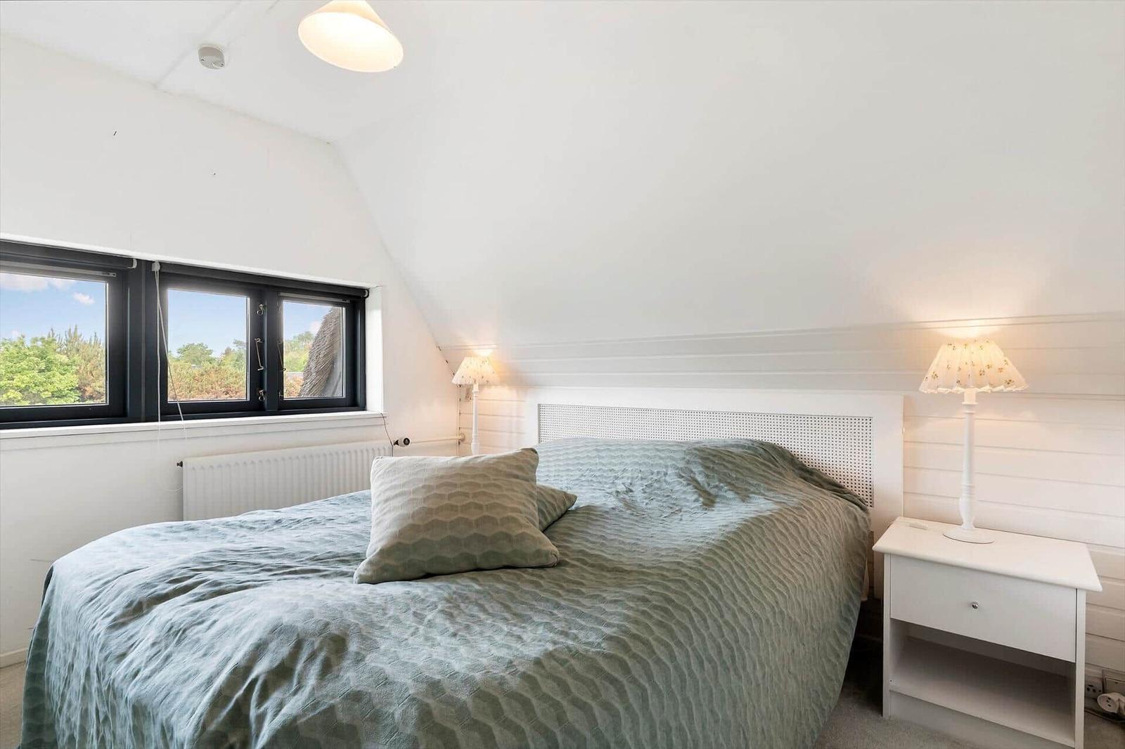 Schlafzimmer mit Bett, Nachttisch und Fenster mit Blick auf Grün.