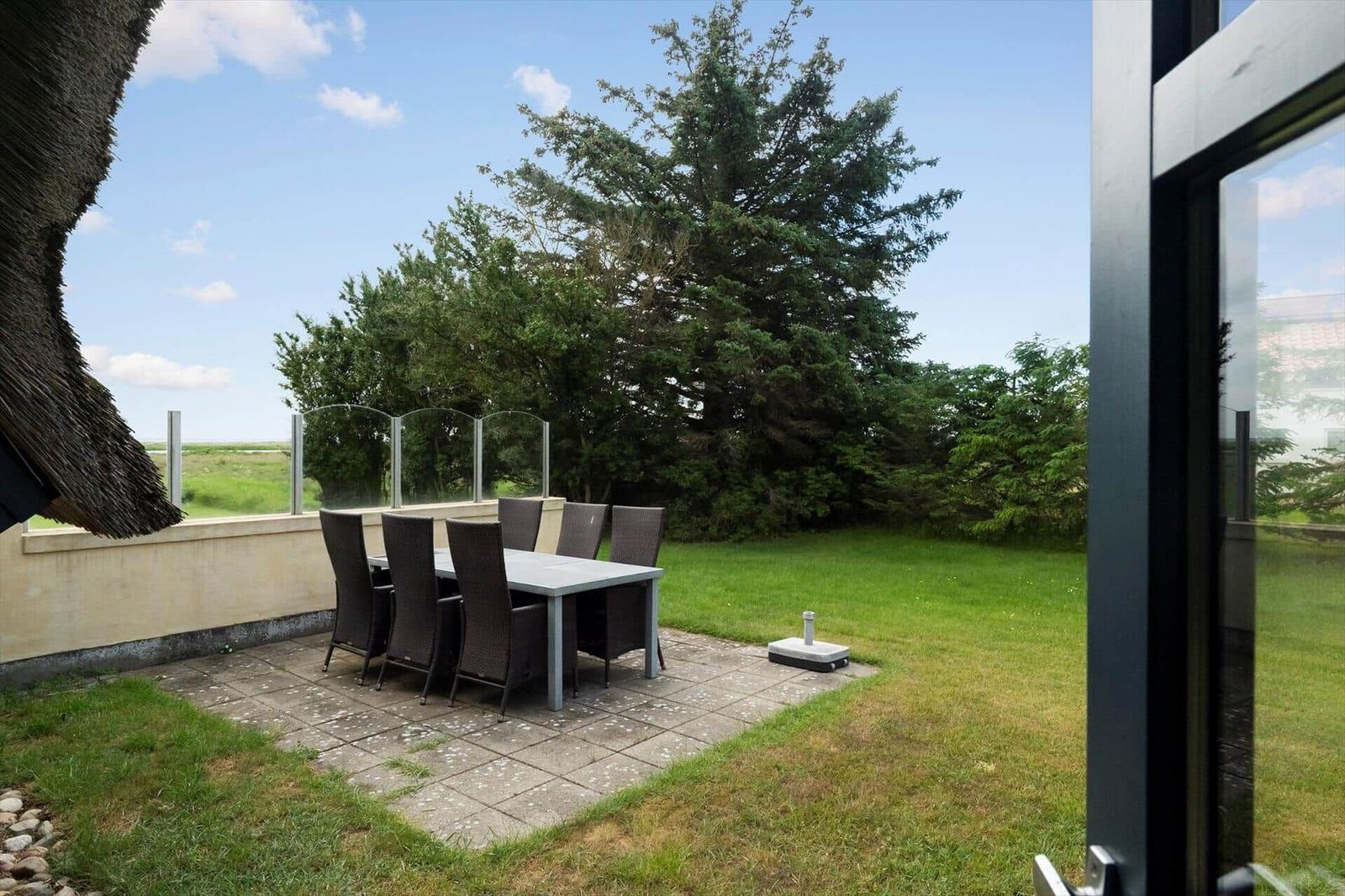 Terrasse mit Tisch und Stühlen, umgeben von Gras und Bäumen unter blauem Himmel.