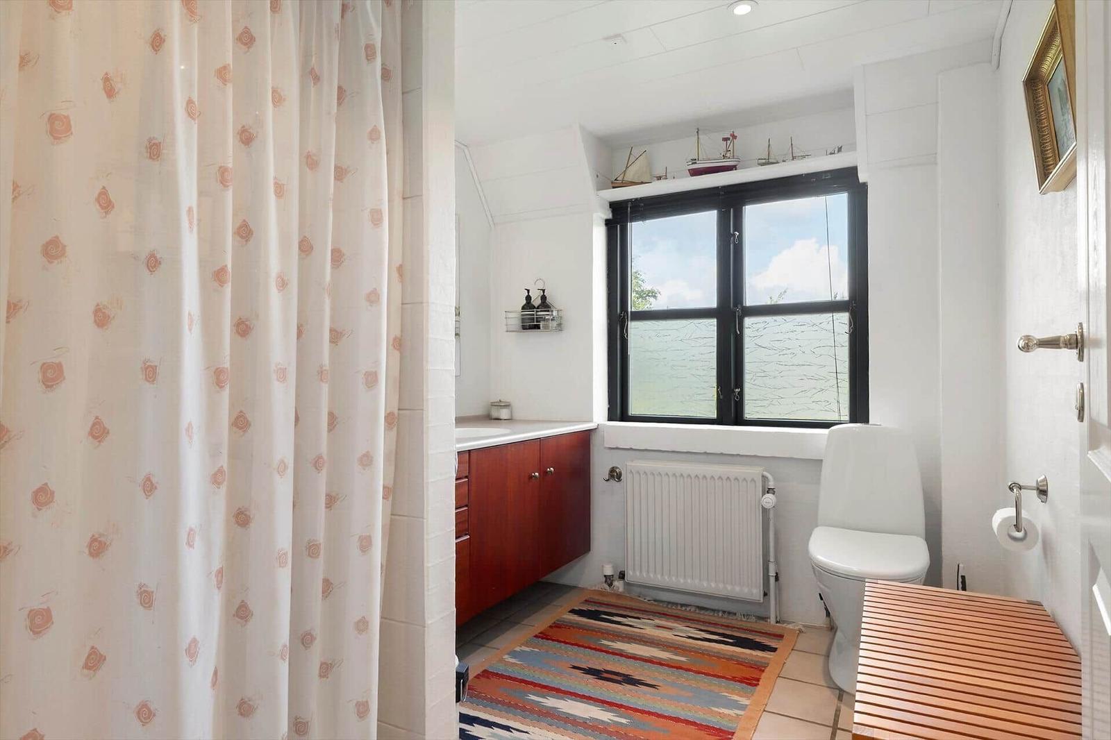Badezimmer mit Fensterblick auf Wasser, Toilette, Holztisch und Badewannenvorhang.