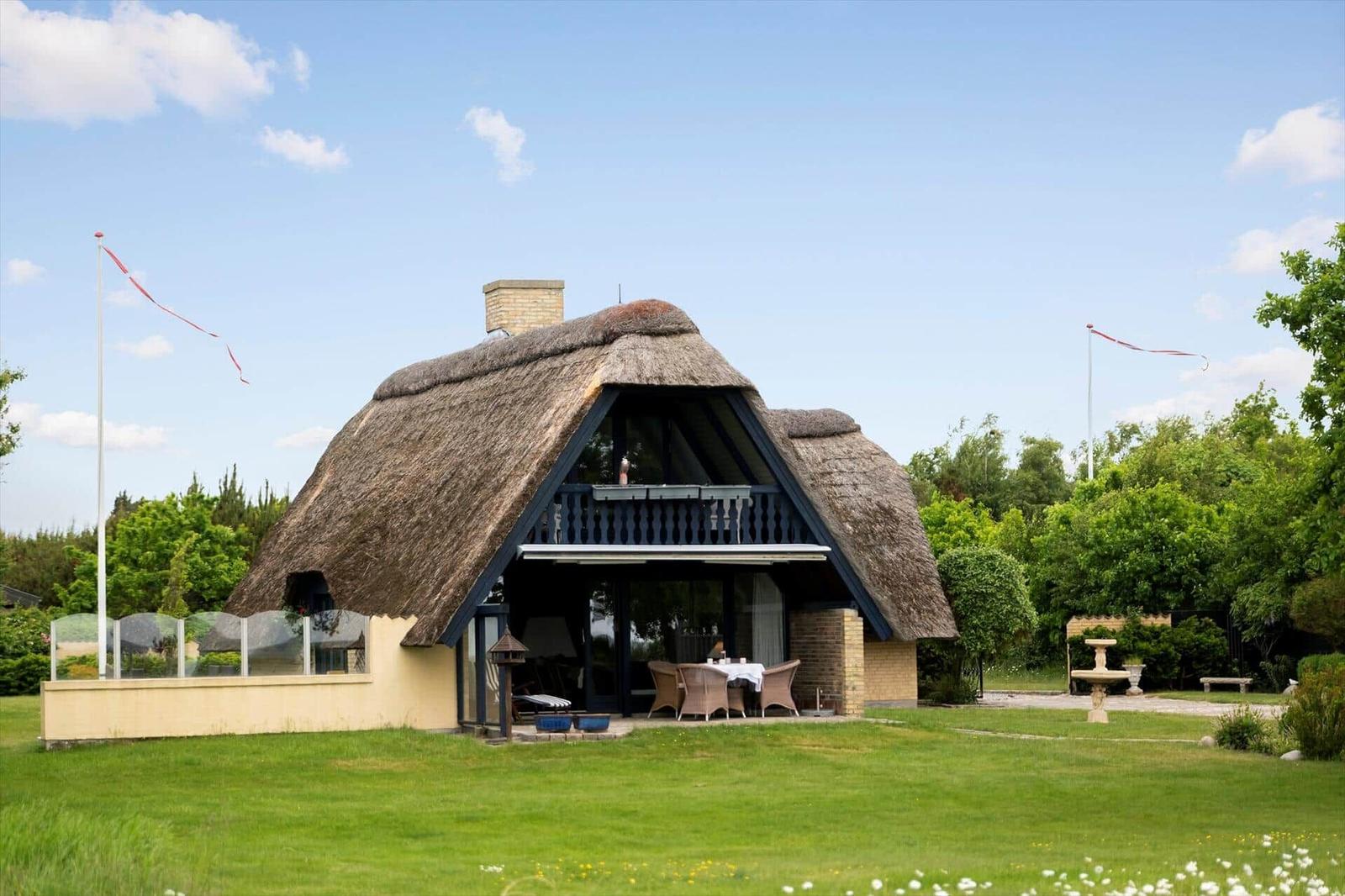 Dorfhaus mit Strohdach, Terrasse und Garten. Zwei Flaggenmasten mit roten Bändern.