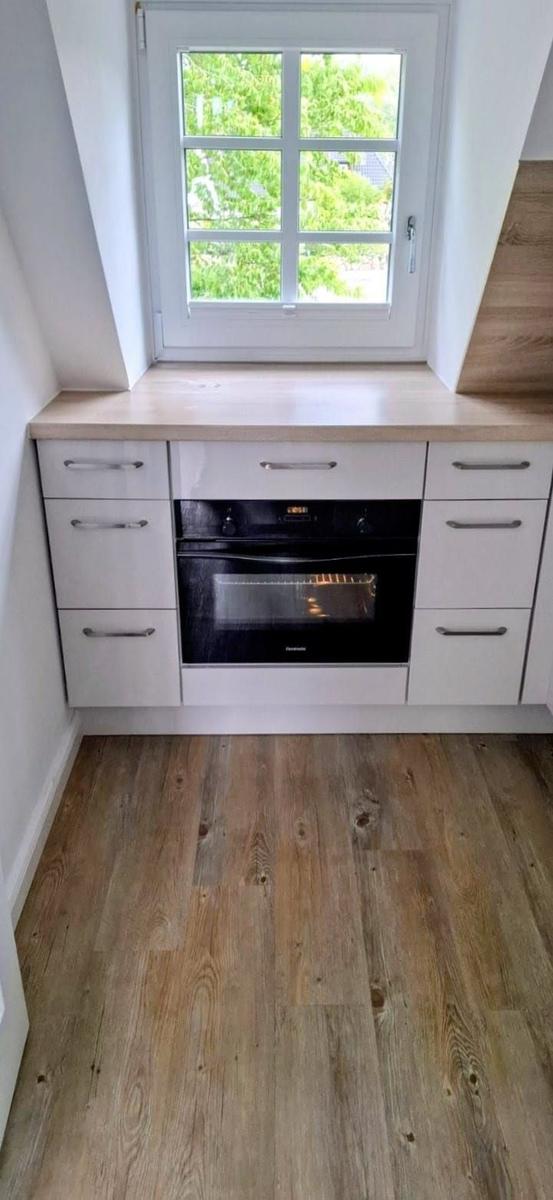 Küchenbereich mit weißer Schrankwand, integriertem Ofen und Holzfußboden unter einem Fenster.