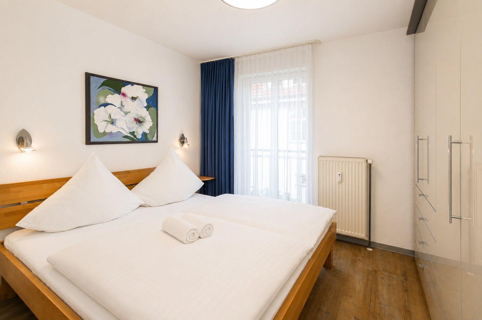 Schlafzimmer mit Doppelbett, Fenster, Gardinen und Kleiderschrank.