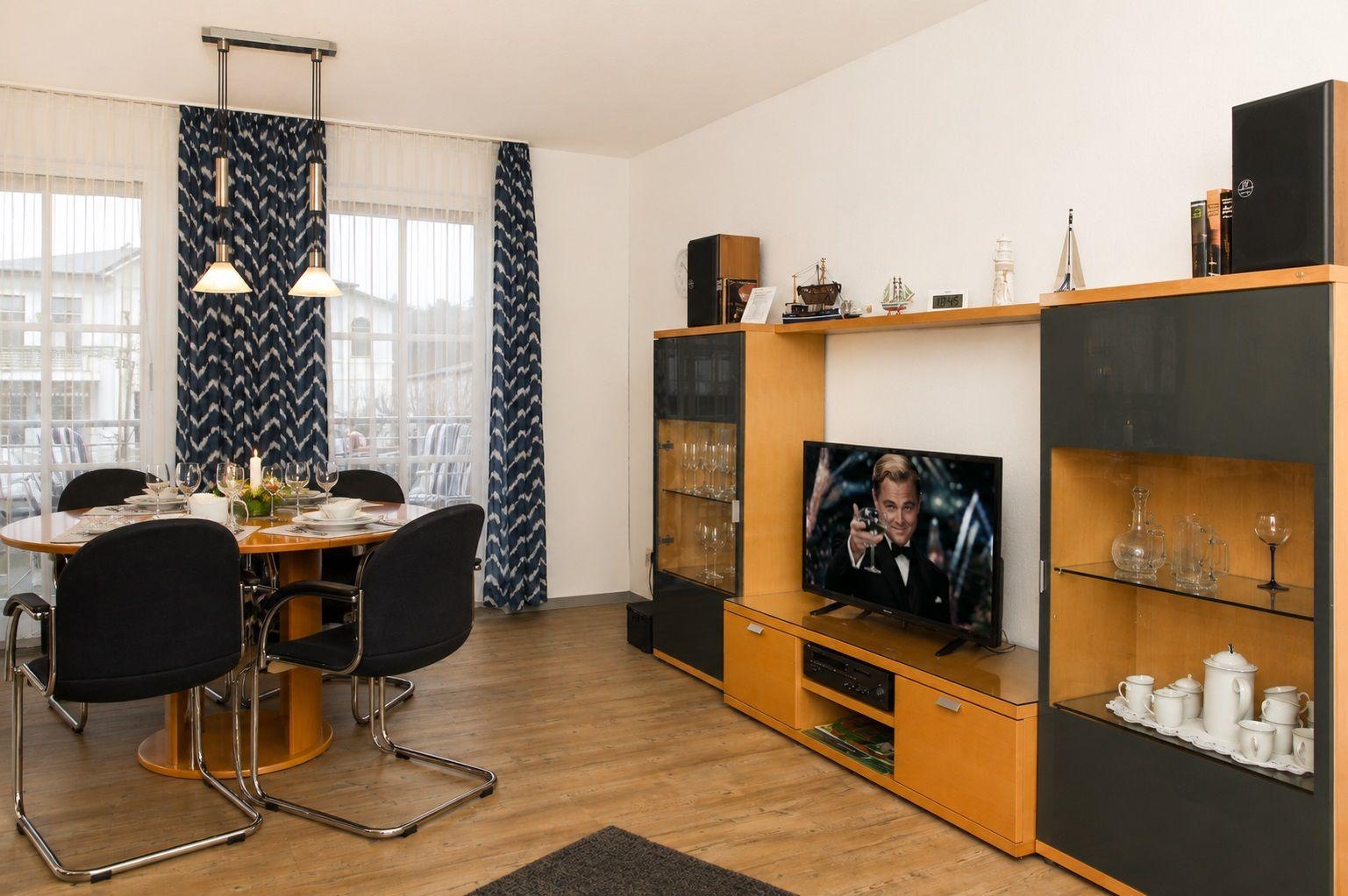Esszimmer mit Tisch, Stühlen und TV-Ecke. Fenster mit Vorhängen. Holzboden.