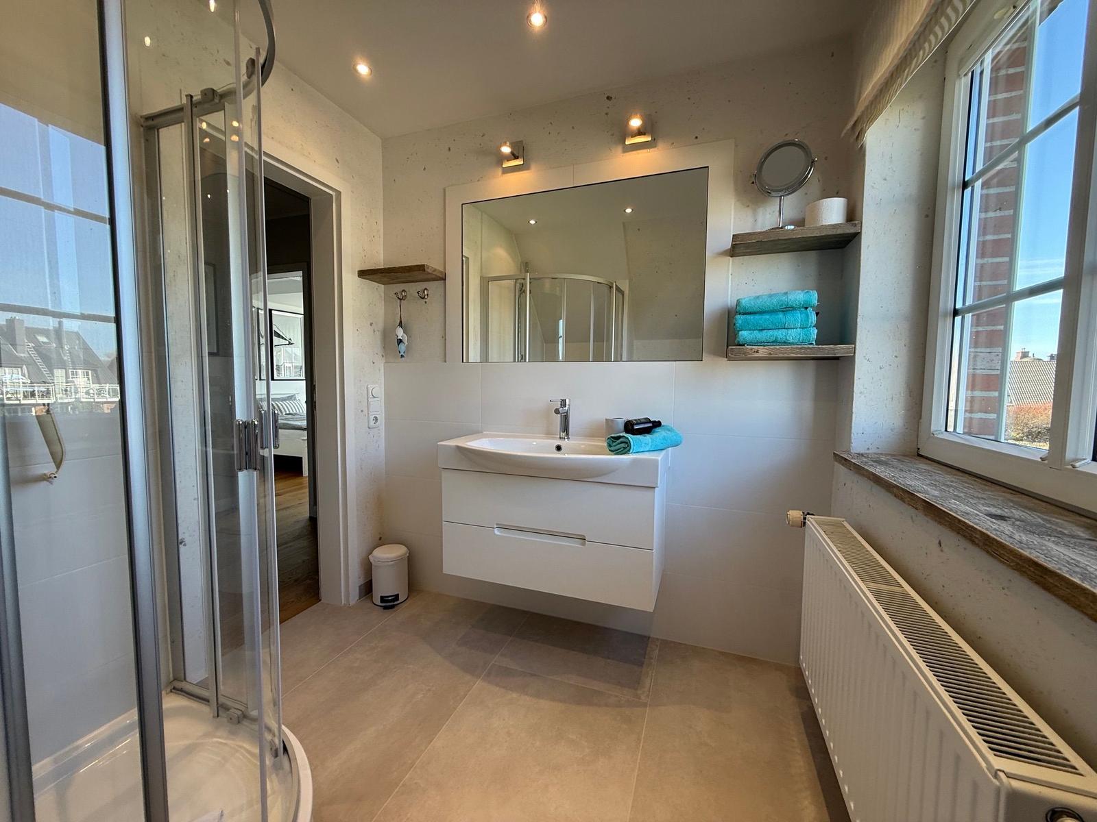 Badezimmer mit Dusche, Waschbecken und Fenster. Auf Wandregal liegen blaue Handtücher.