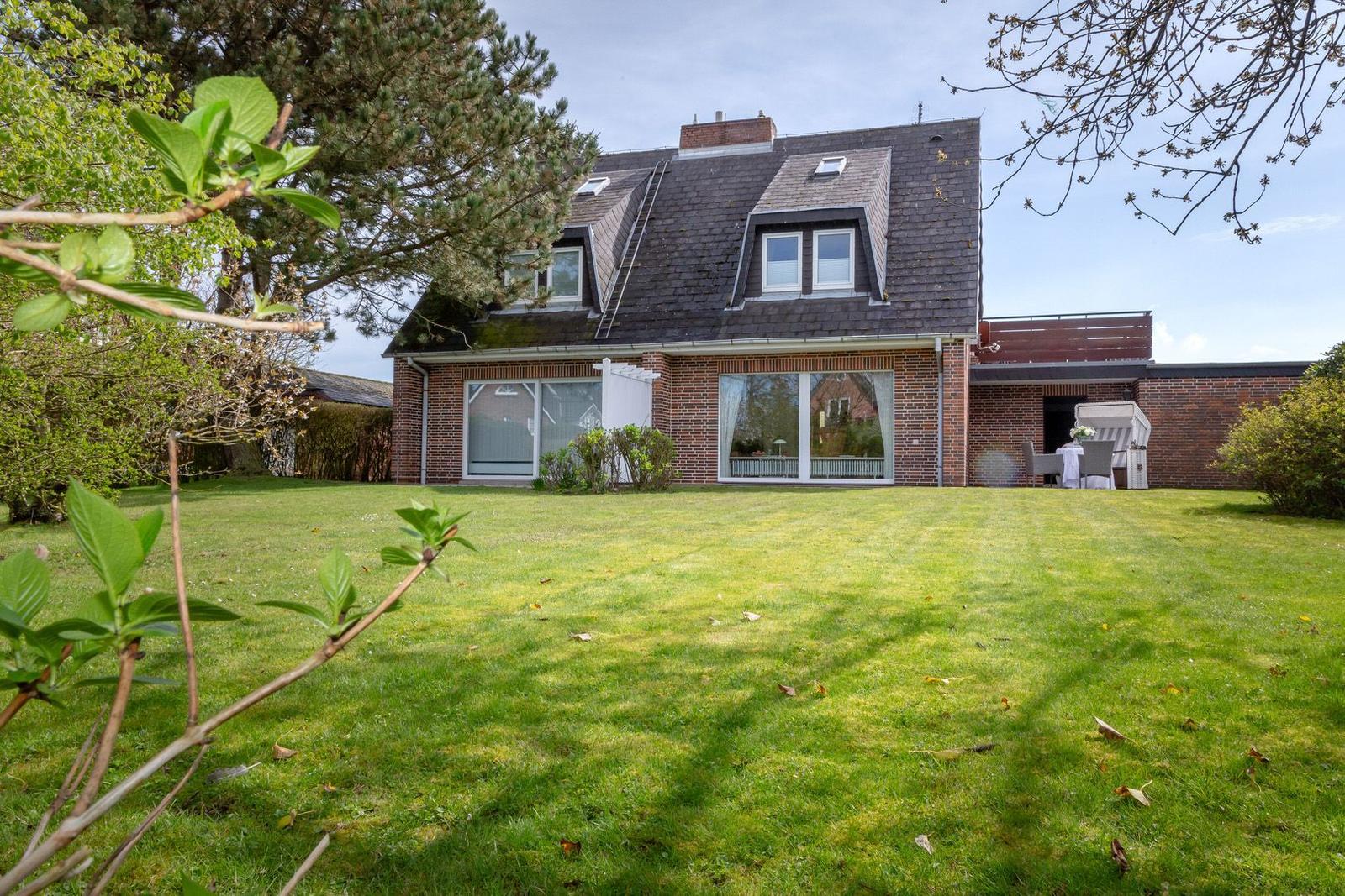 Ein Haus mit großem Garten und Terrasse im Hintergrund. Die Fenster sind groß und hell.