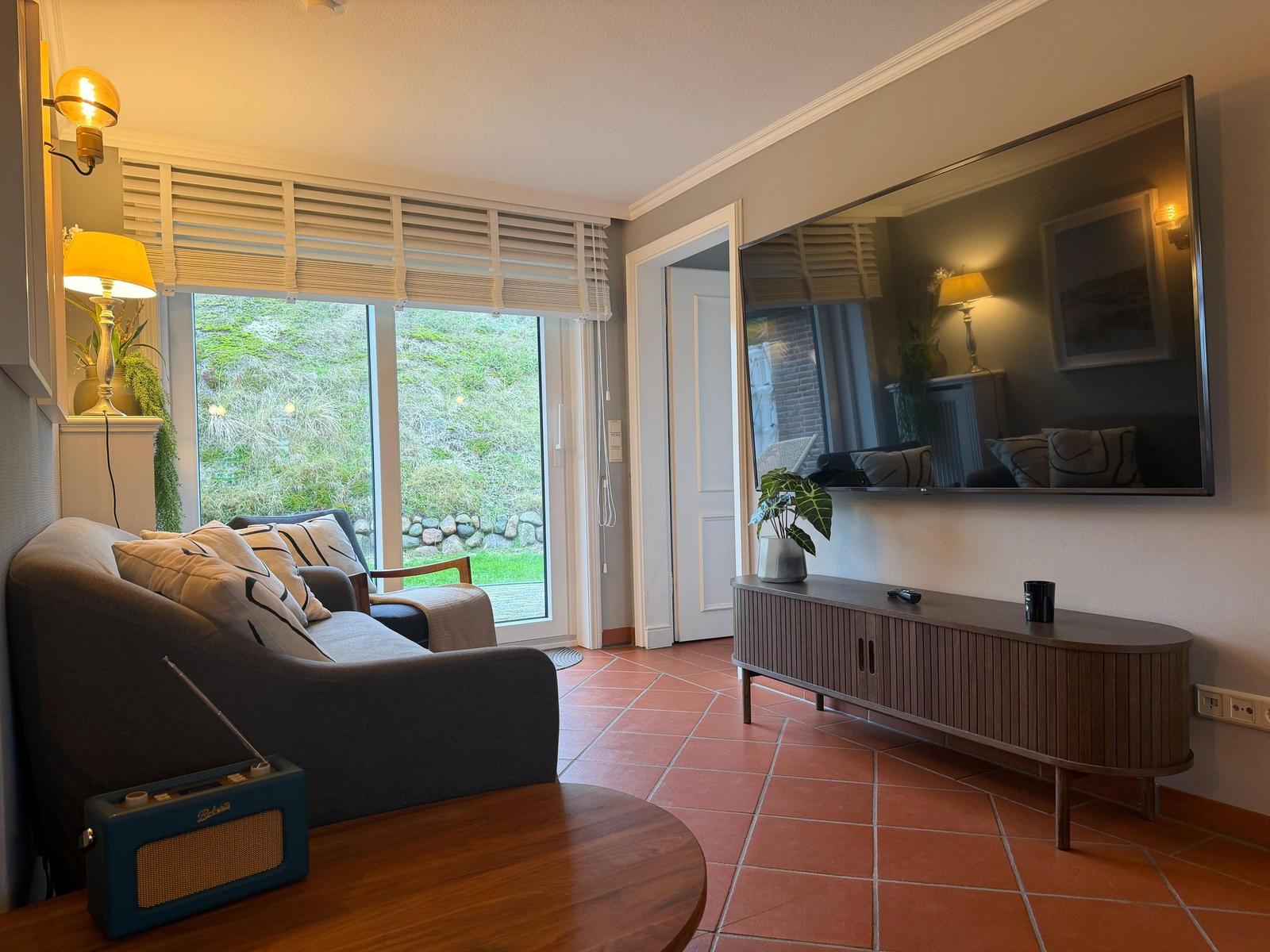 Living room with sofa, TV, and terrace door. Terrace with green area visible.