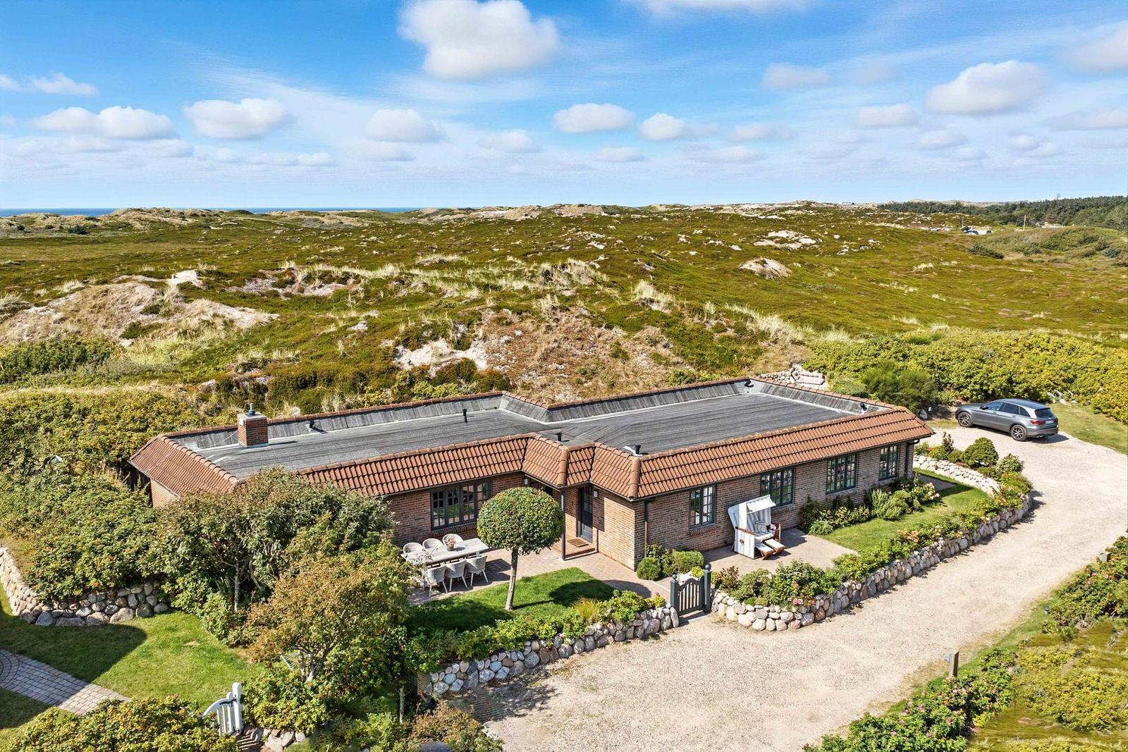 House with terrace and garden, surrounded by dunes and greenery under blue sky.