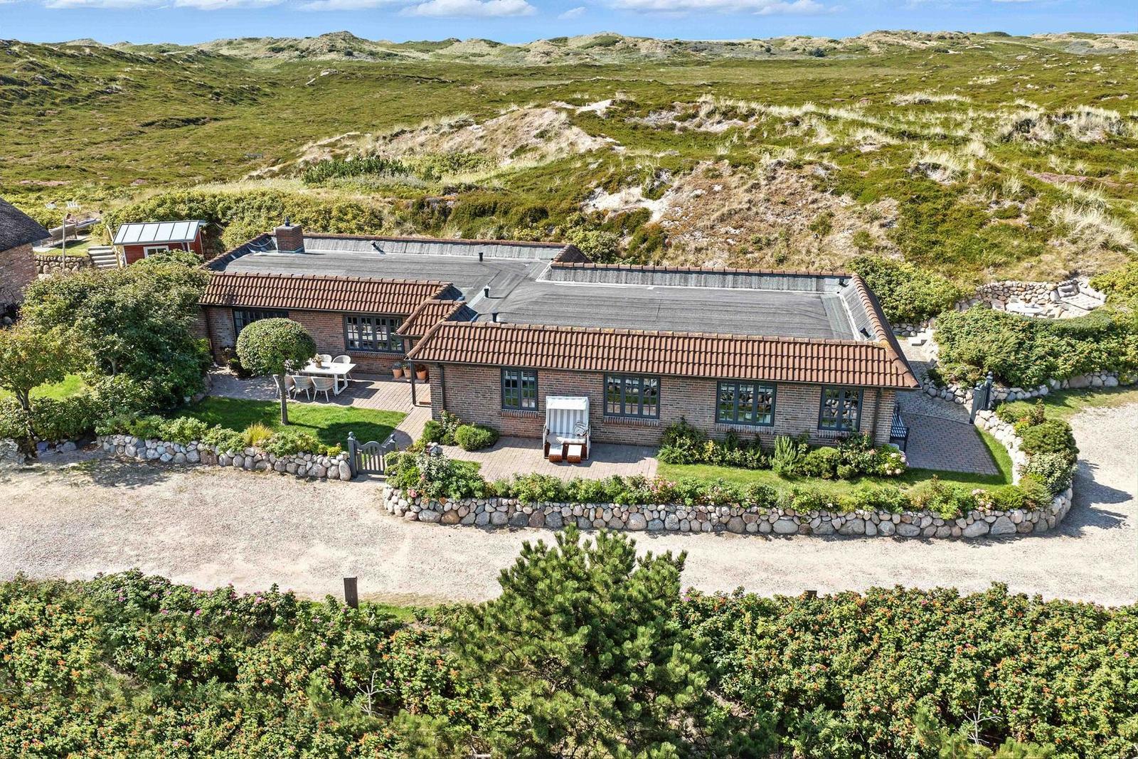 A house with stone walls and terrace is situated in a green, hilly landscape.