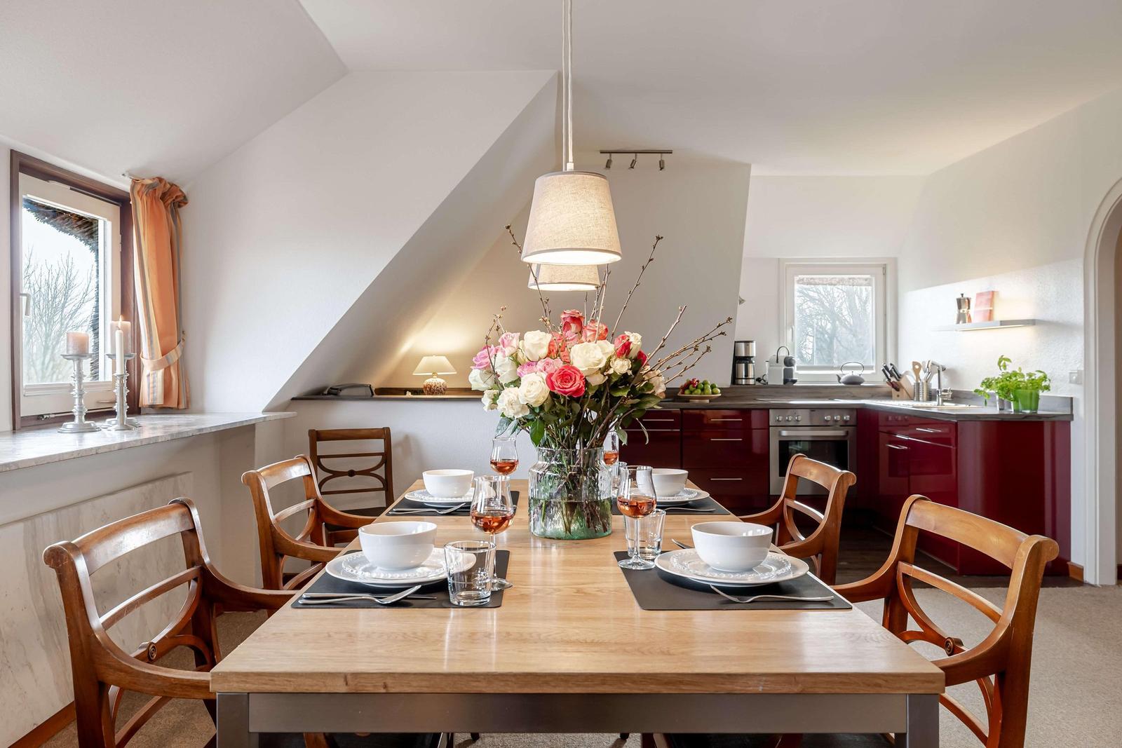 Essbereich mit Holztisch, Stühlen und Blumenvase. Küche mit roten Schränken und Fenster.