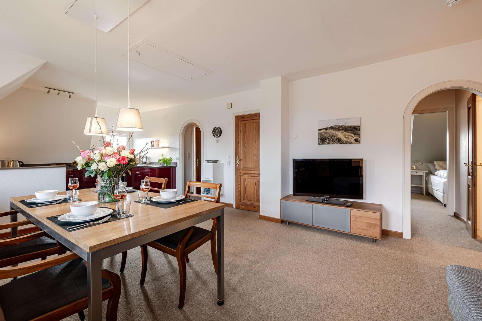 Essbereich mit Tisch, Stühlen und Blumen. Wohnbereich mit TV und Blick in Schlafzimmer.
