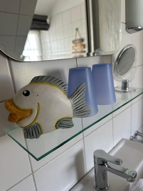 Bad mit Spiegel, Glasregal und Fischdekoration. Zwei blaue Gläser und Waschbecken mit Wasserhahn.