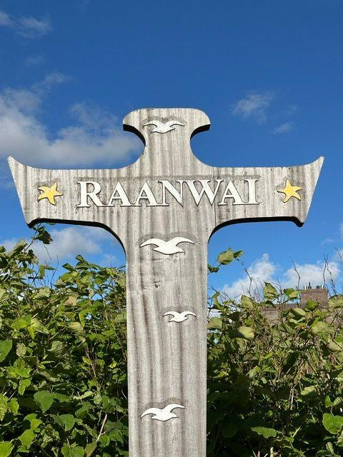 Holzkreuz mit der Aufschrift RAANWAI und stilisierten Möwen. Hintergrund: blauer Himmel und grüne Pflanzen.