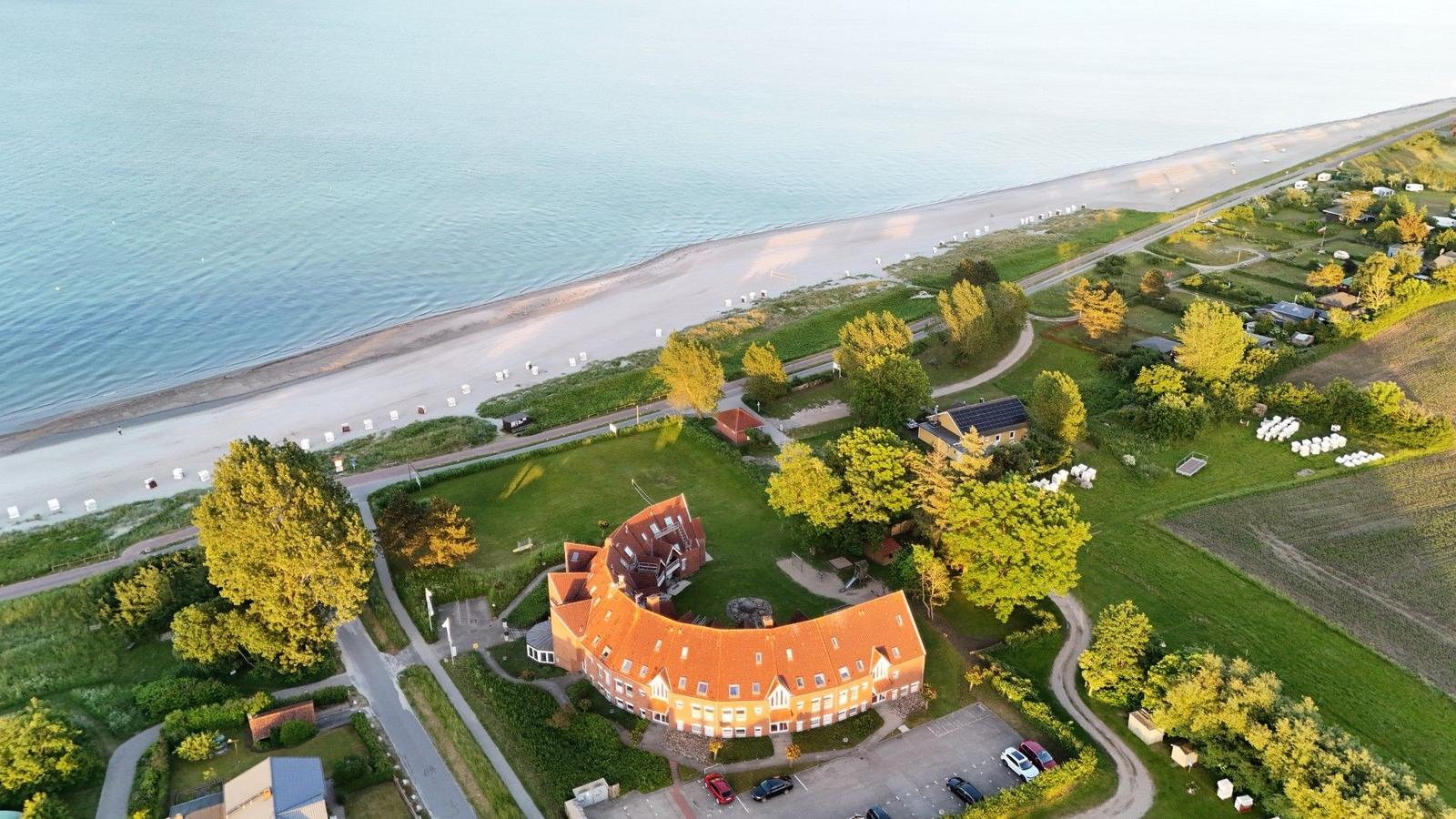 Haus mit rotem Dach liegt am Strand. Grünflächen und Bäume umgeben die Anlage.