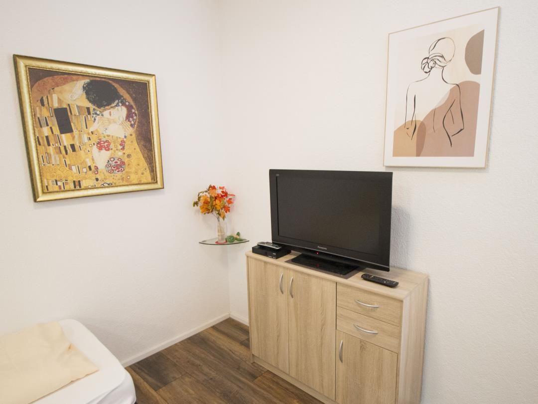 Zimmer mit TV, Schrank, Wandbildern und Blumenvase.